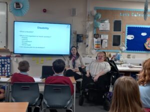 Kat Watkins (DW Staff member), a white woman with blond hair and glasses, wearing a black and white striped shirt and black trousers sat at the front of a classroom in a black and purple electric wheelchair. Next to her is a teacher, a white woman with brown hair wearing a white top, black trousers and a pink and teal scarf. There is a screen next to the teacher with Disability and questions for the learners to answer. There are 4 learners visible two boys on the left with brown hair wearing red jumpers and two girls on the right with dark blond hair.