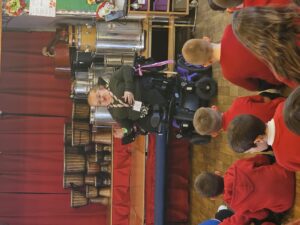 Kat Watkins (DW staff member), a white woman with blond hair wearing glasses, an olive green top and black trousers with a striped lanyard around her neck. She is sitting in a black and purple electric wheelchair. Behind her are lots of drums, in front of her are a group of learners. The learners are all wearing red jumpers. She is talking animatedly. 